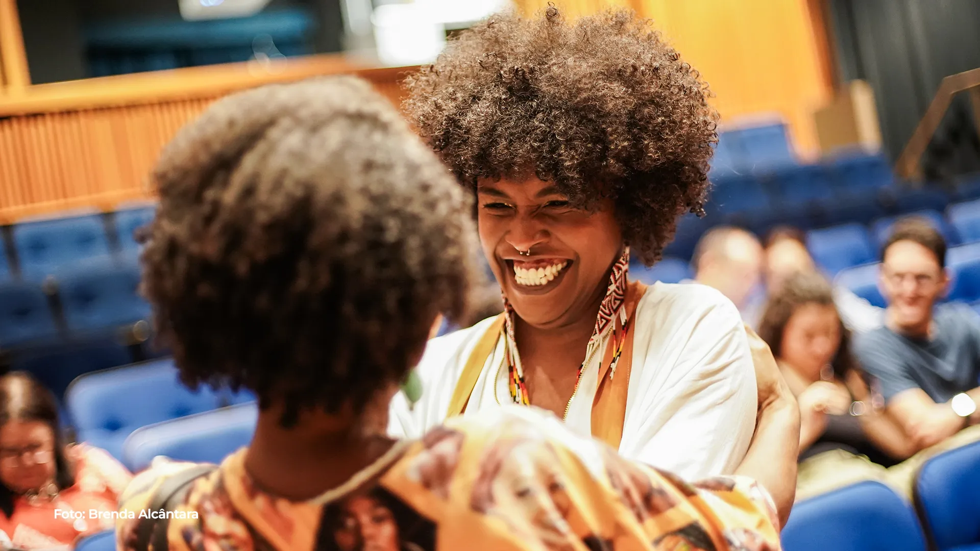 Duas mulheres sorriem lado a lado na plateia