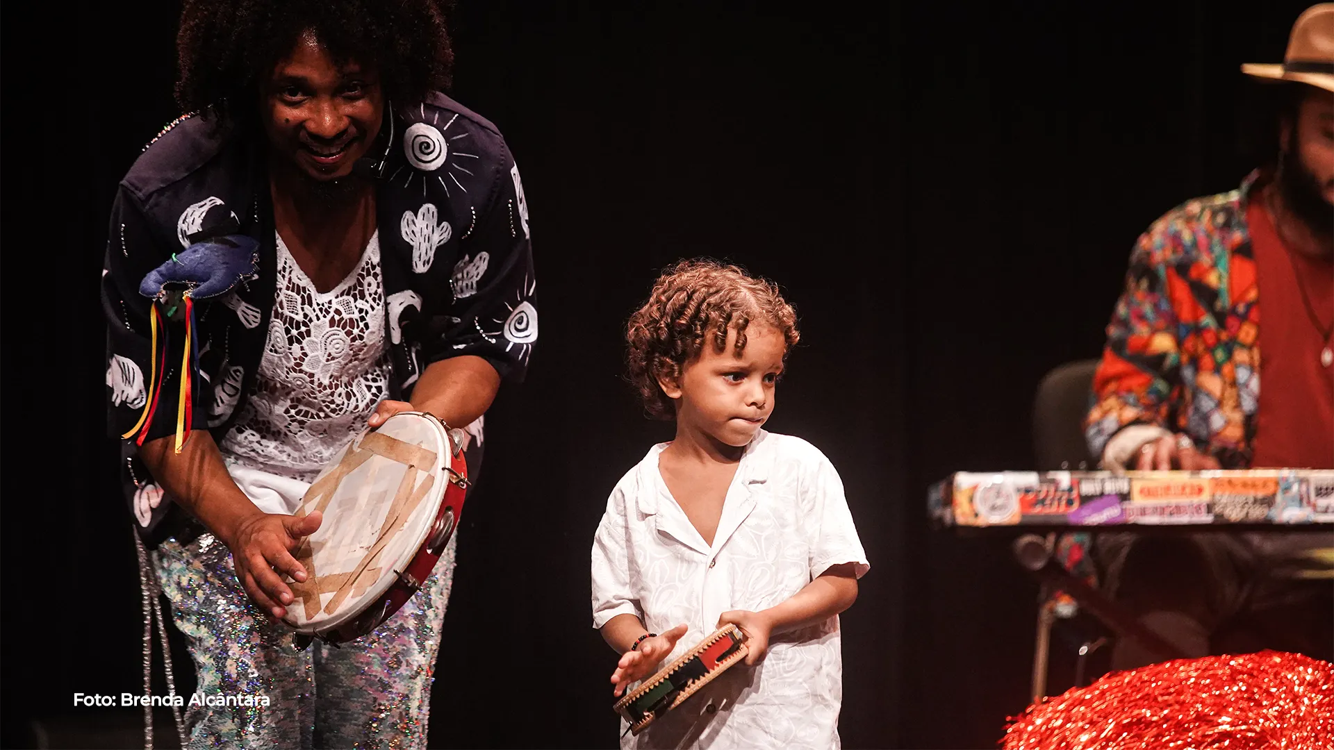 Adulto e criança tocam pandeiro juntos no palco